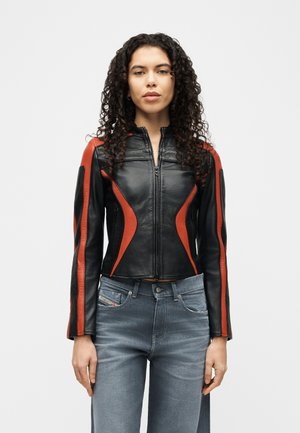 Mujer con cabello rizado que lleva una chaqueta de cuero negra con detalles en rojo y jeans azules, de pie contra un fondo blanco liso.