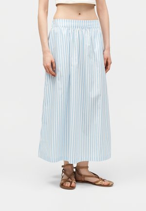 Kvinne iført en midi-skjørt med lyseblå og hvite vertikale striper, en hvit crop top og brune flate sandaler med stropper.