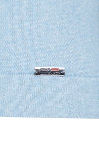 Maglione lavorato a maglia di colore azzurro chiaro con etichetta in metallo argentato con accenti rossi e blu, cucita al centro in basso. Texture morbida.