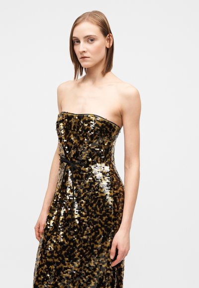 Woman wearing a strapless, sequined gold and black dress with a thin black belt, standing against a plain light gray background.