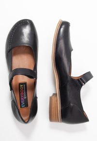 Zapatos Mary Jane de cuero negro con un bajo tacón de madera, correa ajustable, punta cerrada y recortes laterales. Textura suave con detalles de costura.