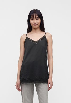 Jeune femme aux longs cheveux foncés portant un débardeur noir bordé de dentelle et un pantalon gris clair, debout devant un fond clair uni.