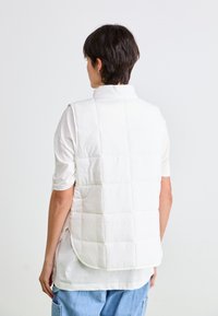 Persoon draagt een witte gewatteerde vest over een wit T-shirt en lichtblauwe jeans, gezien van achteren tegen een effen witte achtergrond.