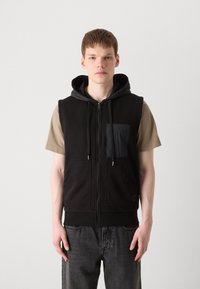 Μαύρο αμάνικο φούτερ με φερμουάρ, με μπροστινή τσέπη, γκρι εσωτερική επένδυση και ραμμένο τελείωμα. Φοριέται πάνω από ένα μπεζ μπλουζάκι και σκούρους τζιν.