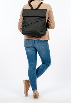 Femme portant un jean bleu, une veste marron clair et des chaussures marron sans lacets, portant un sac à dos carré noir avec une fermeture éclair diagonale à l'arrière.