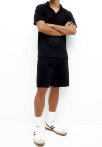 Polo shirt noir en tricot avec un col, porté avec un short noir. Baskets blanches et grises, accompagnées de chaussettes blanches. Modèle debout, les bras croisés.