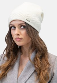 Un bonnet tricoté blanc avec un motif en zigzag texturé, arborant un petit logo en métal. Le matériau semble doux, adapté aux temps froids.