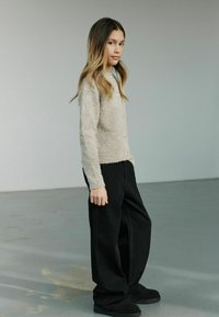 Lys beige strikket cardigan med knapper, parret med brede sorte bukser og mørke slip-on sko. Modellen står sidelæns, hår ned.