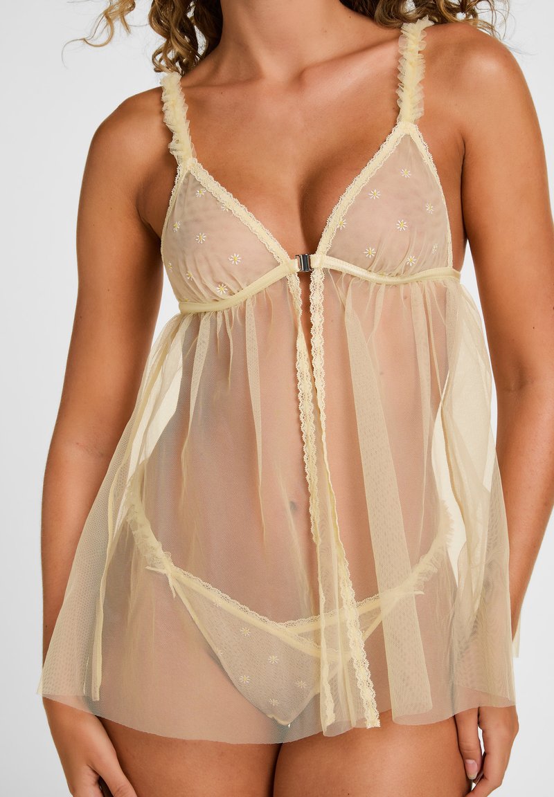 Vrouw draagt doorschijnende lichtgele babydoll lingerie met bloemenborduurwerk en bijpassende slipjes tegen een effen achtergrond.