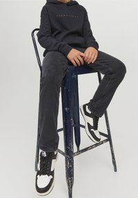 Marineblauer Kapuzenpulli mit orangefarbenem Text, dunkle Jeans und schwarz-weiße Sneaker. Sitzt auf einem abgenutzten blauen Metallhocker.