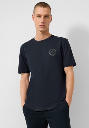 Man met kort haar draagt een donkerblauw t-shirt met een klein rond logo op de linkerborst en staat tegen een eenvoudige lichte muur.
