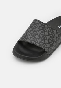 Sandale à talon compensé noire avec une large bride grise ornée d'un motif géométrique sombre et une semelle intérieure texturée sur fond blanc.
