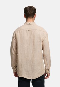 Man wearing long-sleeve beige linen shirt and black pants, standing facing away against a plain white background.