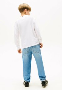 Weißes Langarmhemd mit einem kleinen farbigen Streifen auf der Rückseite, kombiniert mit hellblauen Jeans und schwarzen Schuhen. Lässige Passform und Farbschema.