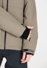 Beige utomhusjacka med dragkedja, strukturerad tyg, svarta ärmslut och en ficka som är markerad med grå accenter. Fitted och funktionell design.