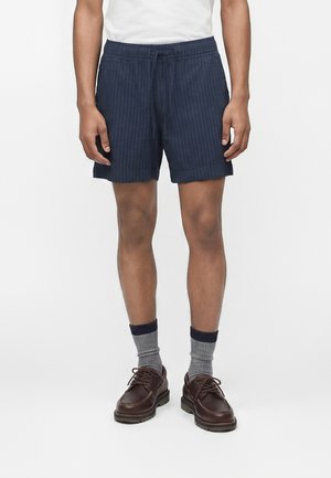 Homme portant un short texturé bleu marine avec cordon, des chaussettes grises bordées de bleu marine, des chaussures en cuir marron et un T-shirt blanc sur un fond blanc uni.