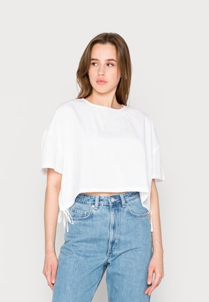 T-shirt cropped de algodão branco com mangas largas, detalhes de cordão na bainha, combinado com jeans de denim azul claro de cintura alta.