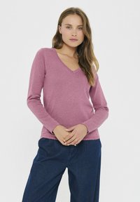 Sieviete ar gariem matiem, tērpta mauve krāsas V-veida kakla garām piedurknēm T-kreklā un brīviem tumši ziliem biksēm, stāv ar rokām saliktām priekšā.