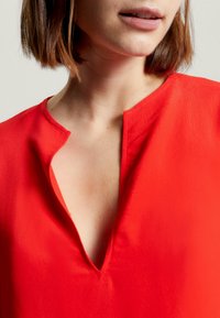 Blusa rossa con scollatura a V, realizzata in un tessuto liscio. Include dettagli di cucitura sottili lungo la scollatura e le spalle.