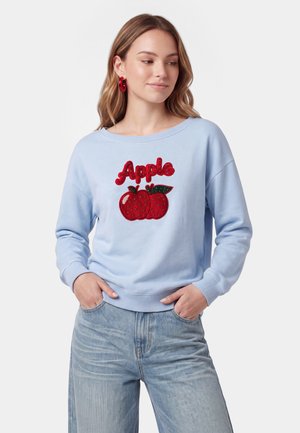 Kvinde iført lyseblå sweatshirt med røde udsmykkede æbler og ordet "Apple," kombineret med lyseblå jeans, hænderne i lommerne.