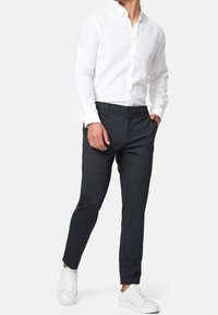 Hombre vistiendo una camisa blanca de botones, pantalones oscuros ajustados y zapatillas blancas, de pie con una mano en el bolsillo.