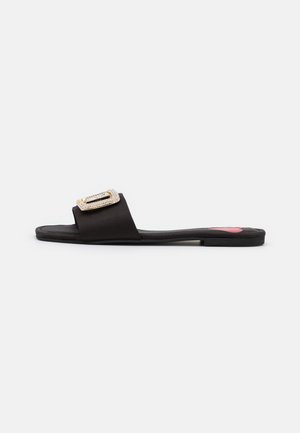 Zwarte platte slipper met een glanzende strap en een rechthoekige strassaccent op de bovenkant. Textuur rubberen zool, minimalistisch ontwerp.