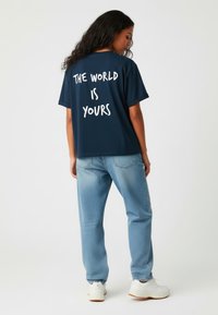 T-shirt bleu marine avec le texte blanc "LE MONDE EST À VOUS" au dos, associé à un jean bleu clair et des baskets blanches. Design à coupe décontractée.