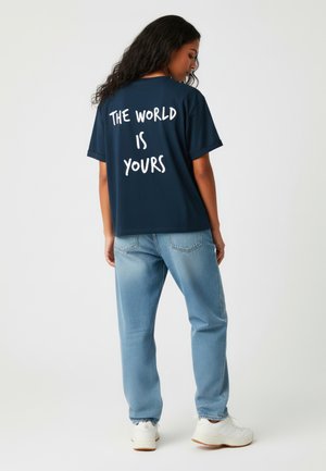 Navyblauwe T-shirt met witte tekst "DE WERELD IS VAN JOU" op de achterkant, gecombineerd met lichtblauwe jeans en witte sneakers. Ontspannen pasvorm ontwerp.
