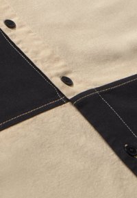 Camisa con cuello que presenta un panel superior beige y un panel inferior negro, con costuras contrastantes y botones negros en el frente.