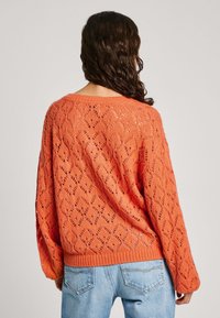 Virkat orange tröja med öppen gallerdesign, ribbat nederkant och lös passform. Kombinerad med ljusblå jeans.