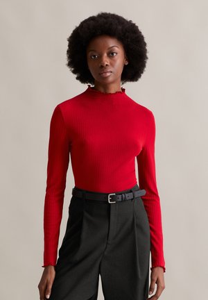 Red ribbed long-sleeve top with a high neck and ruffled trim. Paired with high-waisted black trousers and a black belt.