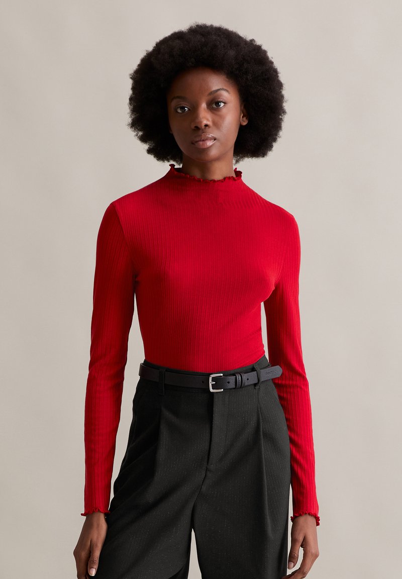 Rotes, geripptes Langarmoberteil mit hohem Kragen und Rüschenbesatz. Kombiniert mit hochgeschnittenen schwarzen Hosen und einem schwarzen Gürtel.