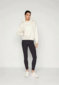 Femeie stând cu o mână în buzunar, îmbrăcată într-un hanorac alb, leggings negri și adidași albi, pe un fundal simplu.