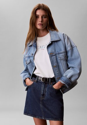 Kvinde iført lyseblå denimjakke over hvid Calvin Klein T-shirt, stukket ind i mørk denim nederdel med sort bælte, hænderne i lommerne.