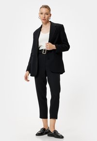 Blazer noir avec épaulettes, top blanc, ceinture noire avec accessoires en or, pantalon noir court et mocassins noirs brillants. Fond uni.