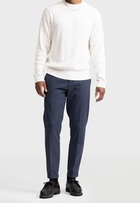 Witte kabelgebreide trui met een ronde hals, gecombineerd met marineblauwe taps toelopende broek en zwarte veterschoenen.