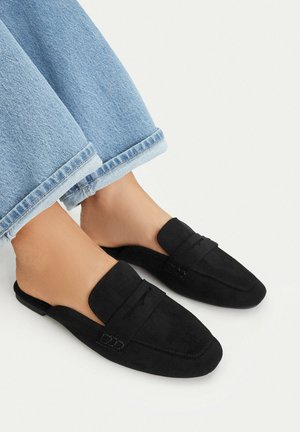 Füße in schwarzen, wildledernen, rückenfreien Loafers, kombiniert mit hellblauen, hochgekrempelten Jeans auf weißem Hintergrund.