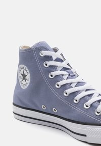 Nieformalny, wysoki niebieski trampek Converse All Star z białymi sznurowadłami, metalowymi oczkami, łatką z logo na kostce i białą gumową podeszwą z czarnymi paskami.