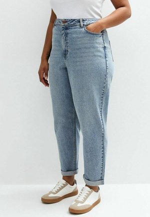 Persona con jeans azul claro remangados, zapatillas blancas con suelas marrones y una camisa blanca metida, de pie con una mano en el bolsillo.