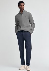 Maglione grigio a maglia con colletto a metà zip, abbinato a pantaloni chino blu navy e sneakers chiare. Il maglione ha polsini a coste e un dettaglio con logo.