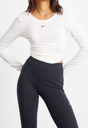 Mujer vistiendo una camiseta corta de manga larga blanca y leggings deportivos negros de cintura alta, ambos con el logo Swoosh de Nike.