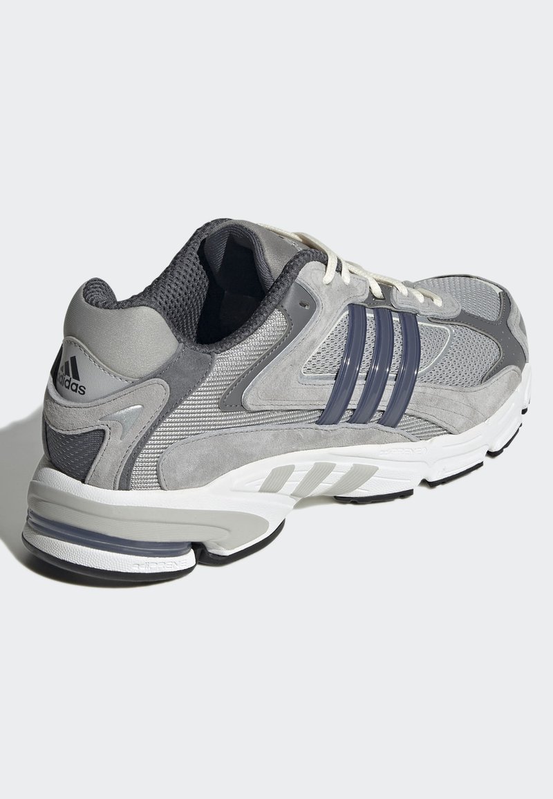 Zapatilla deportiva gris con materiales de ante y malla, con tres franjas azules, suela blanca gruesa y un talón acolchado para mayor soporte.