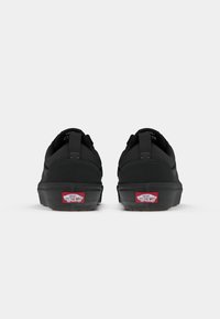 Svarte Vans-sko har et strømlinjeformet design med teksturert canvas, polstrede krager og røde logo-detaljer på hælen.