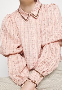 Blouse rose clair avec un motif texturé, manches bouffantes et col festonné. Présente des accents marron et des motifs floraux délicats.