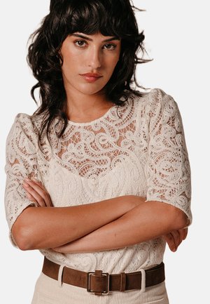 Donna con capelli scuri e ondulati che indossa una maglia color crema in pizzo con maniche a sbuffo, braccia incrociate e una cintura marrone sopra pantaloni beige.