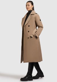 Cappotto beige con design doppiopetto, grande colletto, bottoni neri e vita cintrata, abbinato a leggings neri e stivali.