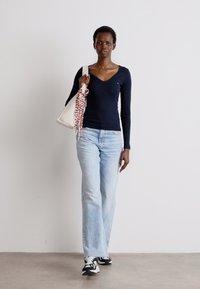 Tricou bleumarin cu mânecă lungă și decolteu în V, blugi deschiși la culoare cu talie evazată, geantă albă cu un model roșu, adidași alb-negru, stând pe un fundal neutr.