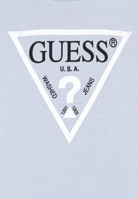 Világoskék póló, fehér háromszög alakú logóval, fekete szöveggel, amely tartalmazza a "GUESS", "USA", "WASHED" és "JEANS" feliratokat, valamint egy középső kérdőjelet.