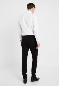 Man i vit skjorta, svarta slim-fit byxor och svarta skor som står vänd bort från kameran mot en neutral bakgrund.