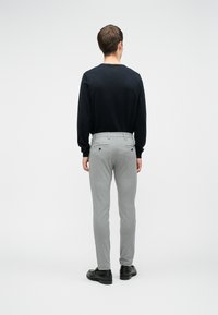 Homme debout de dos portant une chemise noire à manches longues, un pantalon gris clair avec poches arrière et des chaussures habillées noires sur fond blanc.